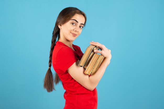 Jonge vrouwelijke student in rood t-shirt met boeken die zich op blauwe muur bevinden.