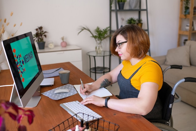 Jonge vrouwelijke student notities maken in Kladblok zittend achter computermonitor thuis en scherm kijken tijdens webinar
