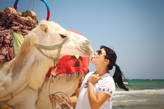 Jonge vrouwelijke toerist in tuniek en zonnebril poses met kamelen tegen achtergrond Middellandse Zee, zandstrand. Meisje op vakantie. Spelen met kamelen. Badplaats in Tunesië, Afrika, vroege lente