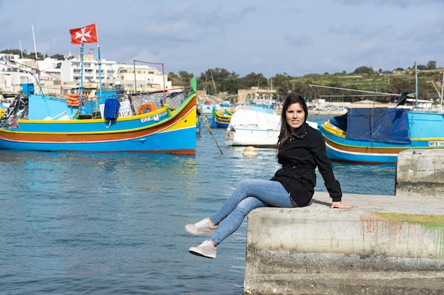 Jonge vrouwelijke toerist zit en op de achtergrond de zee vol kleurrijke boten bij Marsaxlokk in Malta