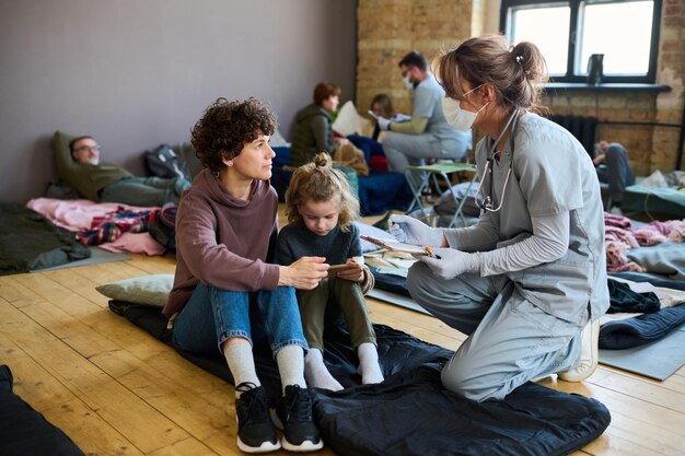 Jonge vrouwelijke vrijwilliger in medisch uniform die vluchteling met kind raadpleegt