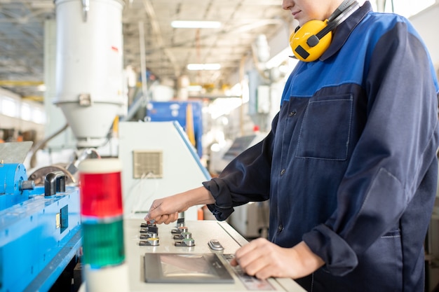 Jonge vrouwelijke werknemer in overall die voor het bedieningspaneel van de industriële machine staat en aan knoppen draait terwijl hij deze voorbereidt op het werk