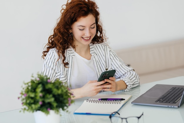 Jonge vrouwelijke werknemer zittend op het bureau en het gebruik van mobiele telefoon