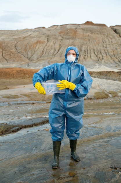 Jonge vrouwelijke wetenschapper in beschermende conalls, gasmasker, rubberen handschoenen en laarzen met plastic container met water- en grondmonsters