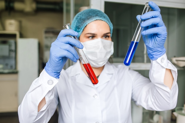 Jonge vrouwelijke wetenschapper met medische pet en beschermend masker doet onderzoek met reageerbuizen