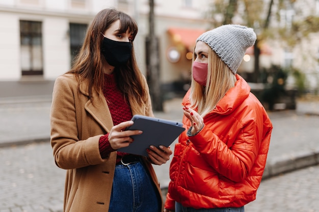 Jonge vrouwen in medische maskers die zich bij stadsstraat bevinden en digitale tablet houden. Vriendinnen tijd buitenshuis doorbrengen met modern apparaat.