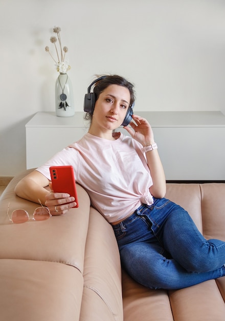 Jonge vrouwen luisteren naar muziek met koptelefoon met mobiele telefoon in de hand zittend op de beige bank thuis