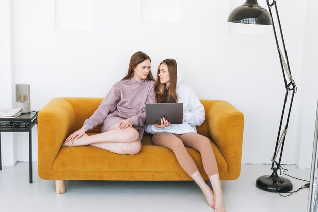 Jonge vrouwen zussen tweeling tieners in hoodie mooie meisjes met behulp van laptop zittend op gele bank