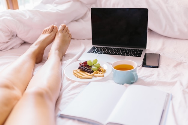 Jonge vrouwenbenen. Thee thuis in haar bed, haar laptop nakijken, een boek lezen en ontbijten.