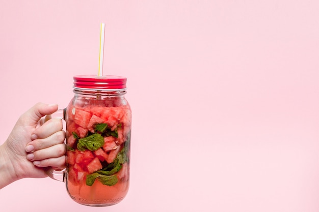 Jonge vrouwenhanden die vers gedrukte watermeloenlimonade van citrusvruchten houden. vrouw met metselaar pot vol koude cocktail, muntblaadjes. roze achtergrond, kopie ruimte, close-up