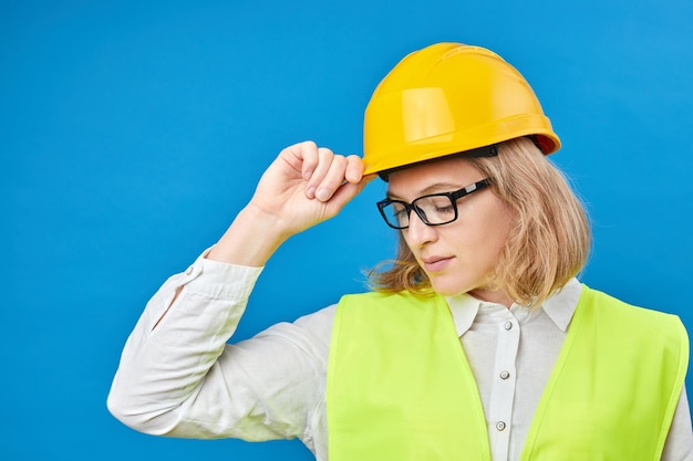 Jonge vrouweningenieur die een gele helm draagt die zich in studio op blauwe achtergrond bevindt Het concept van de decoratie van de bouwreparatie Studio schieten