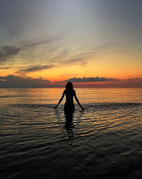 jonge vrouwensilhouet bij oranje zonsondergang in de zeewatergolf en roze bewolkte hemel