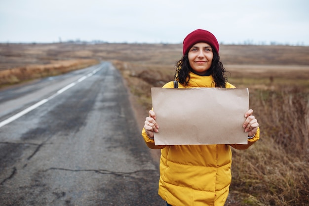 Jonge vrouwentoerist die gele jas en rode hoed draagt, staat met een blanco posterpapier aan de kant van een lege winterweg.
