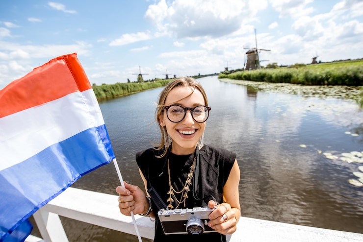 Foto jonge vrouwentoerist die zich met nederlandse vlag op het mooie landschap backgorund met oude windmolens in nederland bevindt