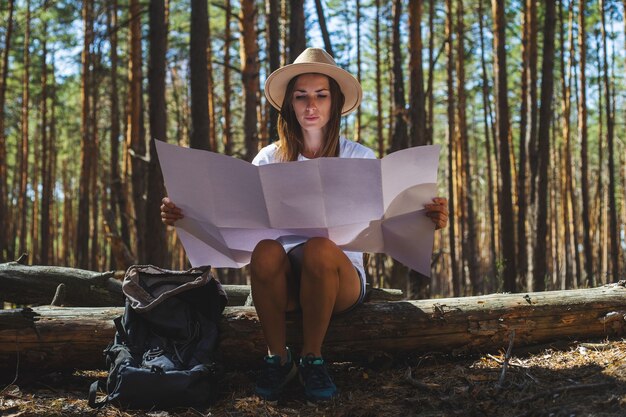 Jonge vrouwentoerist in een hoed en een t-shirt zit op een logboek en kijkt naar een kaart tijdens een pauze in het bos.