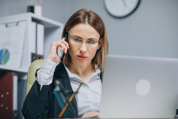 Jonge vrouwenzitting bij bureau gebruikend laptop en sprekend op de telefoon