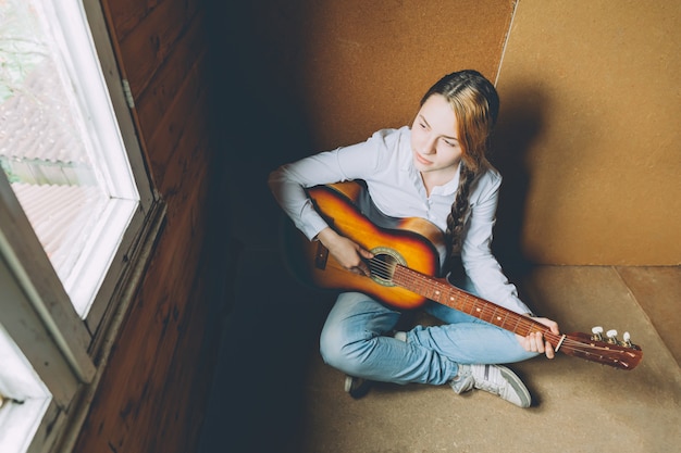 Jonge vrouwenzitting in ruimte op vloer en thuis het spelen van gitaar.