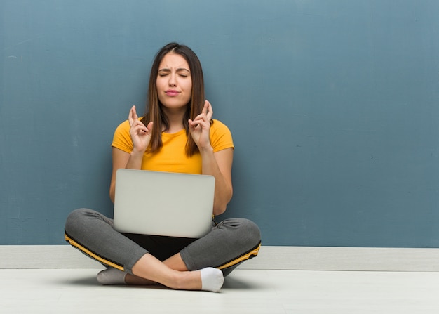 Jonge vrouwenzitting op de vloer met laptop die vingers kruisen voor het hebben van geluk