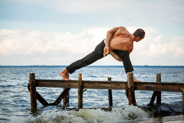 Foto jonge yogatrainer die parivrita parswakonasana beoefent, poseert op een houten pier aan een zee- of rivieroever