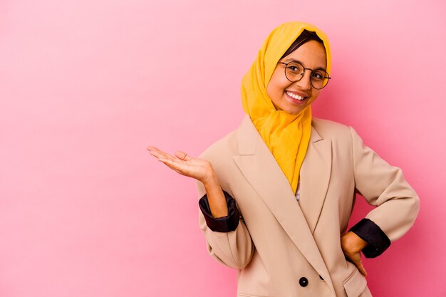 Jonge zakelijke moslimvrouw die op roze muur wordt geïsoleerd die een exemplaarruimte op een palm toont en een andere hand op taille houdt