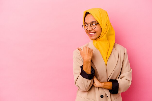 Jonge zakelijke moslimvrouw geïsoleerd op roze achtergrond wijst met duimvinger weg, lachend en zorgeloos.