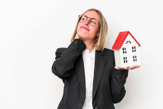 Jonge zakenkaukasische vrouw die een speelgoedhuis vasthoudt dat op een witte achtergrond wordt geïsoleerd en achterhoofd aanraakt en een keuze maakt