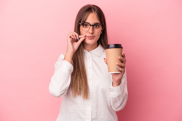 Jonge zakenkaukasische vrouw die een take-away houdt geïsoleerd op roze achtergrond met vingers op lippen die een geheim houden.