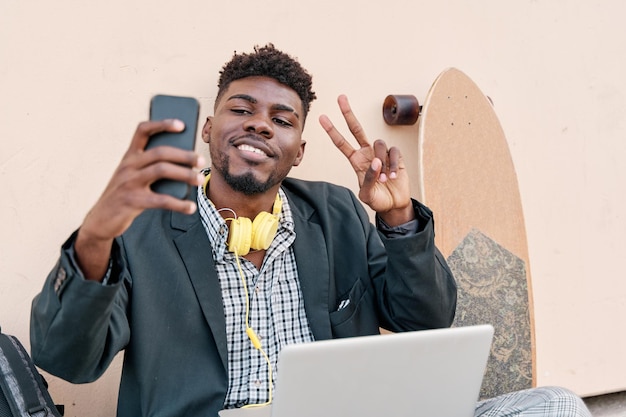 Jonge zakenman die buiten zit en een selfie maakt met zijn smartphone terwijl hij op zijn laptop werkt