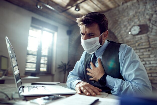 Jonge zakenman die een gezichtsmasker draagt en pijn op de borst voelt tijdens het werken op een laptop op kantoor tijdens een virusepidemie