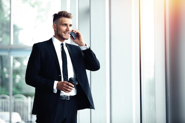 Jonge zakenman die in een kantoor bij het raam staat en op een mobiele telefoon praat