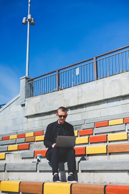 Jonge zakenman die online werkt met een laptop terwijl hij op een bankje buiten zijn kantoorgebouw zit