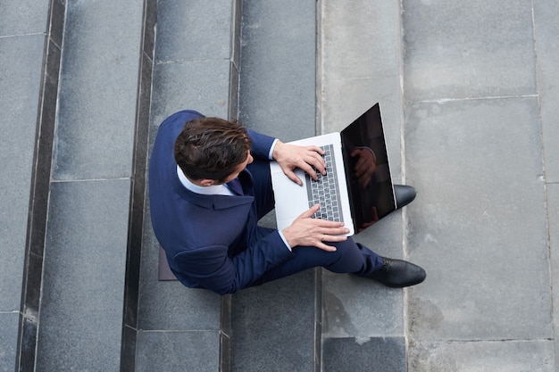 Jonge zakenman die op moderne laptop werkt en buiten op de trap zit, bovenaanzicht