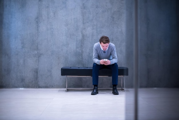 jonge zakenman die slimme telefoon gebruikt terwijl hij tijdens een pauze op de bank in de kantoorlobby zit