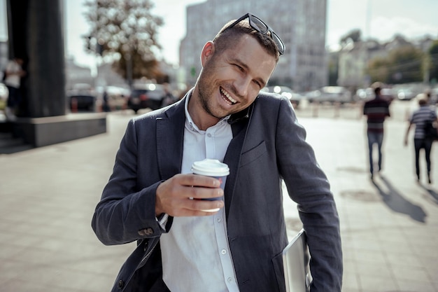 Foto jonge zakenman die zijn laptop vasthoudt terwijl hij zijn telefoon gebruikt en koffie drinkt. op straat staan.