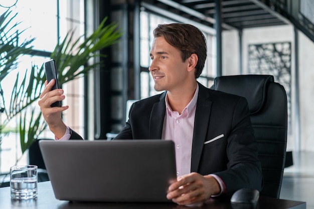 Jonge zakenman glimlachend kijken naar de telefoon dragen zwart pak zittend aan tafel met laptop Officemanager met telefoon werken in modern kantoor met glazen ramen wazig achtergrond