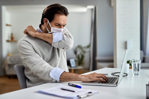 Jonge zakenman hoest in elleboog terwijl hij een beschermend gezichtsmasker draagt en thuis op een computer werkt