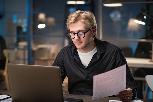 Jonge zakenman in bril die laptop gebruikt om een contract te typen terwijl hij tot laat in de nacht op kantoor op zijn werkplek werkt