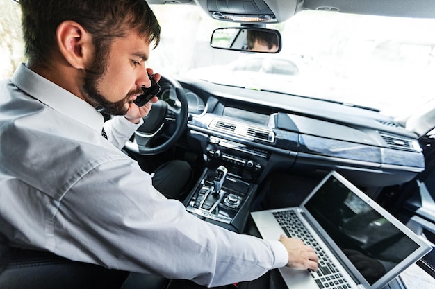 Jonge zakenman in pak bij auto met mobiel en laptop