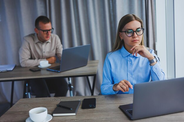 Jonge zakenman in vrijetijdskleding zit aan een bureau en lacht met een digitale tablet terwijl collega's op de achtergrond werken in een modern, ruim kantoor