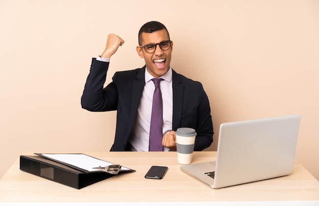 Jonge zakenman in zijn kantoor met een laptop en andere documenten die een overwinning vieren