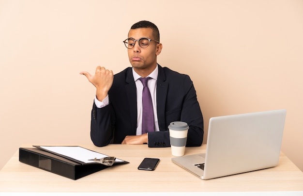 Jonge zakenman in zijn kantoor met een laptop en andere documenten ongelukkig en wijst naar de kant