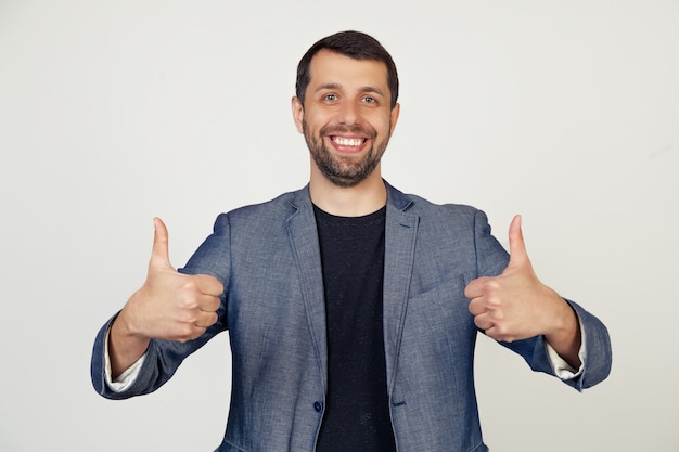 Jonge zakenman man met een baard in een jas, een teken van succes maakt een positief gebaar met zijn hand, duimschroef opwaarts glimlachend en blij.