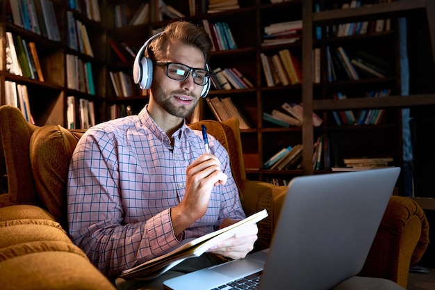 Foto jonge zakenman mannelijke student die koptelefoon draagt e-learning op laptop computer zit in een stoel werkt vanuit thuis kantoor online leren studeren op afstand training cursus webinar stream kijken