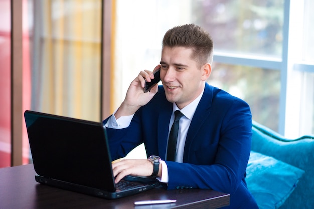 Jonge zakenman met behulp van laptopcomputer tijdens een gesprek op mobiele telefoon in een modern kantoor