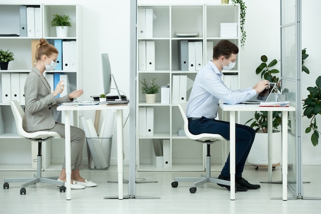 Jonge zakenman met beschermend masker en formalwear die laptopscherm afveegt met nat doekje terwijl zijn collega ontsmettingsmiddel op handen spuit