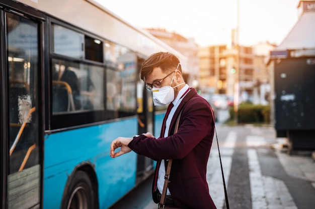 Jonge zakenman met beschermend masker staat alleen op lege straat en wacht op bus of taxi. Virus pandemie of vervuiling concept.