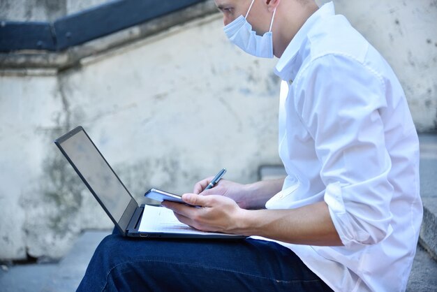 Jonge zakenman met masker die buiten op laptop werkt