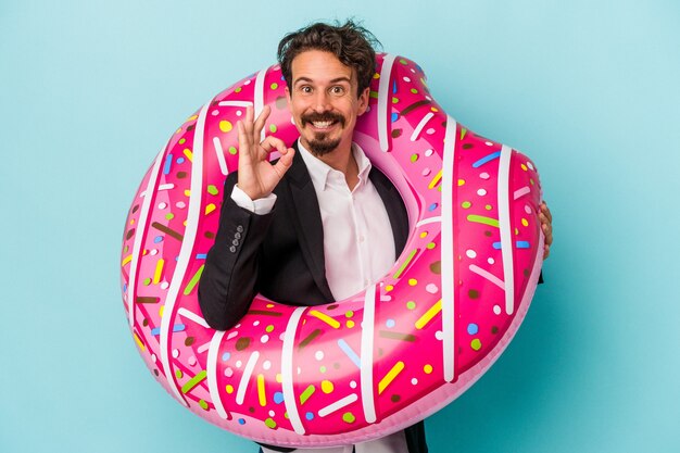 Jonge zakenman met opblaasbare donut geïsoleerd op blauwe achtergrond vrolijk en zelfverzekerd weergegeven: ok gebaar.