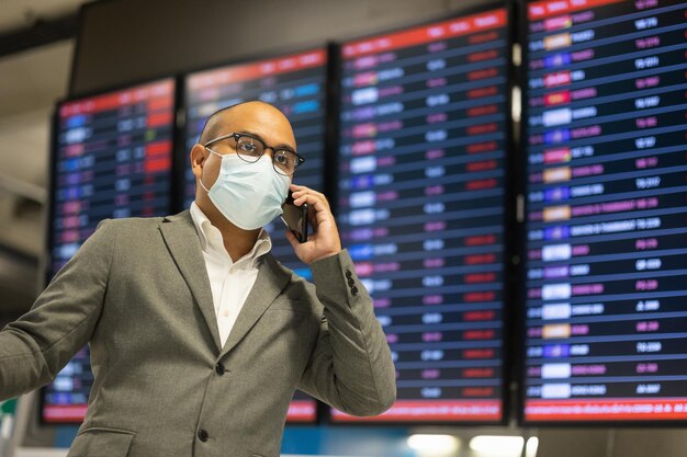 Jonge zakenman praten over de telefoon op de luchthaven. Masker dragen. reizen tijdens een pandemie moet social distancing zijn. Hij staat bij het wachtende vliegtuig van het informatievluchtschema op de luchthaventerminal.