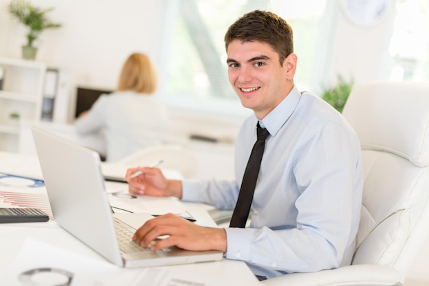 Jonge zakenman zit aan een bureau voor een laptop en werkt. camera kijken.